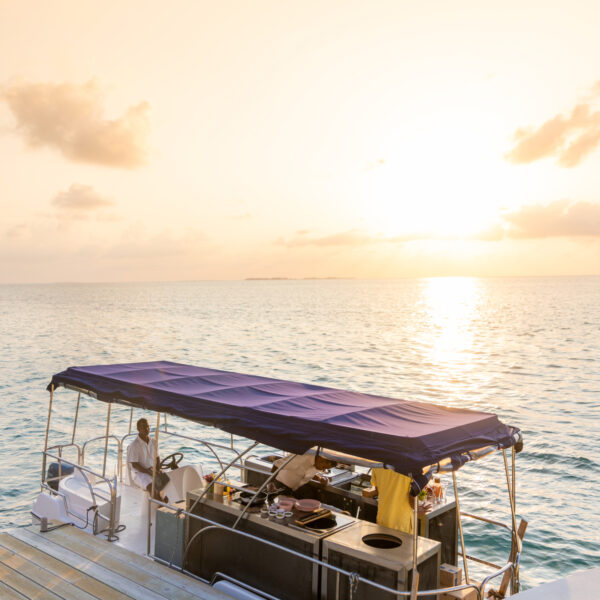 A boat with a purple canopy is docked at a wooden pier during sunset. Two people are on the boat, one seated at the helm and the other standing near a counter. The calm ocean reflects the golden hues of the setting sun, creating a serene atmosphere—perfect for an exclusive Soneva Maldives experience.