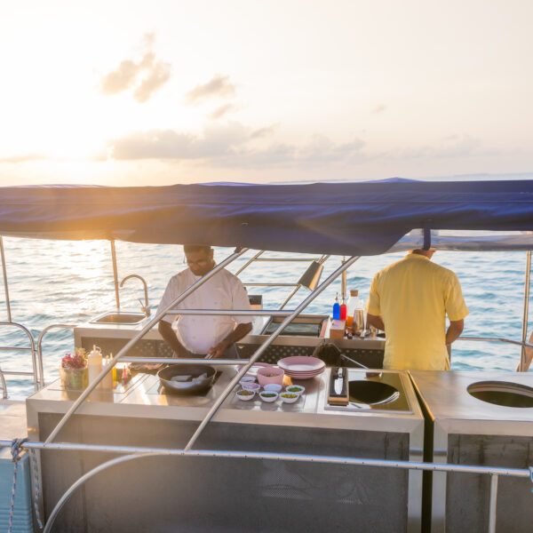 Two people stand on a boat at sunset under a canvas canopy, working at an outdoor kitchen setup. Various cooking utensils and ingredients are laid out in front of them, with the ocean and a setting sun in the background—a true Maldives exclusive experience by Soneva.