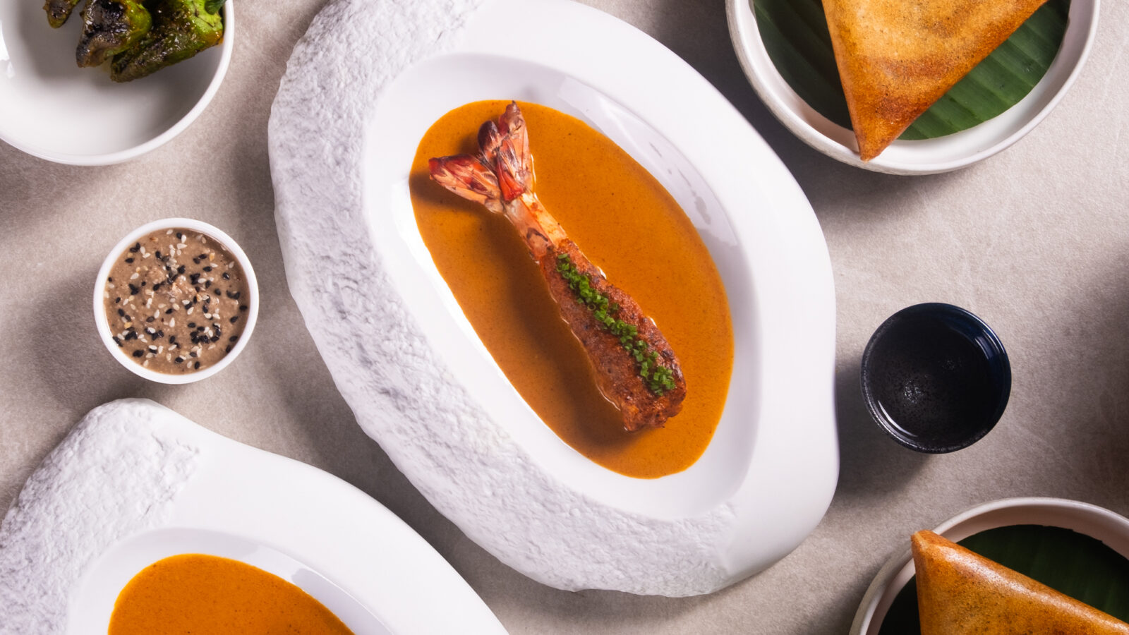 An elegant food presentation featuring two dishes of soup with garnished seafood, triangular pastries on leaves, small bowls of sauce and spices, roasted peppers, and a glass of green beverage. The setting is on a light stone surface.