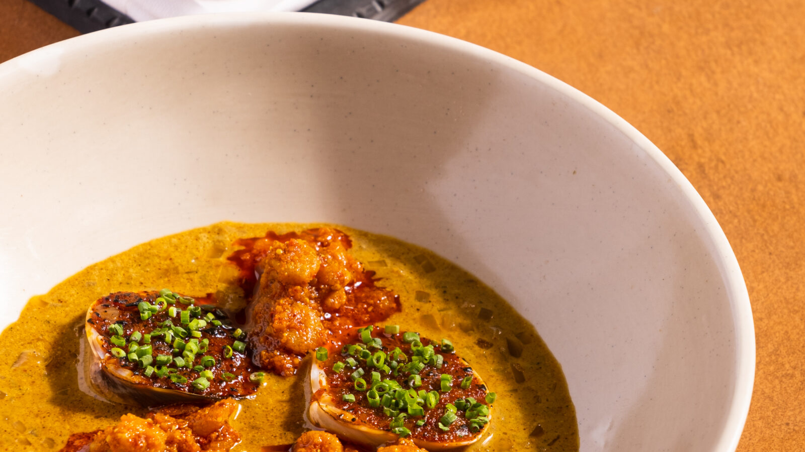 A white bowl filled with a savory dish featuring scallops topped with a chunky sauce and chopped chives, set in a creamy sauce. In the background, golden-brown bread rolls on a black tray and white paper are visible on a wooden surface.