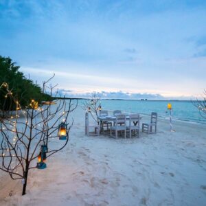 A serene beach setup at the Maldives Luxury Resort features a dining table and chairs on the sand, surrounded by leafless trees adorned with string lights and lanterns. The calm ocean is visible in the background, with a light blue sky merging with the horizon as evening approaches, promising an exquisite Soneva dining experience.