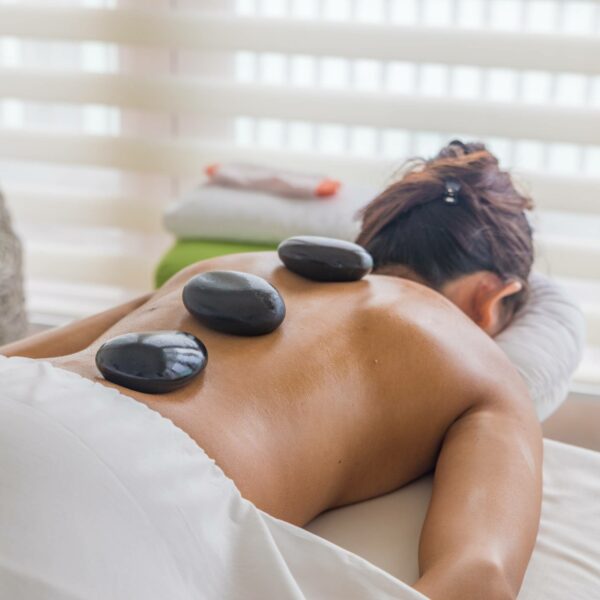 A person is lying face down on a Maldives spa massage table with three large hot stones placed on their bare back. They are covered with a white towel from the waist down. The background features soft lighting and blurred window blinds, creating a calming atmosphere perfect for wellness and relaxation.