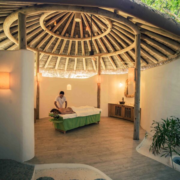 A serene Maldives spa scene inside a circular hut with a thatched roof. A person lies on a massage table, covered with a green sheet, receiving a massage from a therapist. The space has natural light, white walls, wooden accents, and potted plants, creating an ideal wellness retreat.