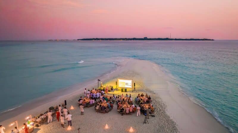 Aerial view of a group gathering at a small sandy beach extension surrounded by the calm ocean waters of Soneva Maldives at sunset. People are seated around tables with a central stage featuring a large screen. Soft lighting and decor create a luxury festive atmosphere. An island is visible in the background.