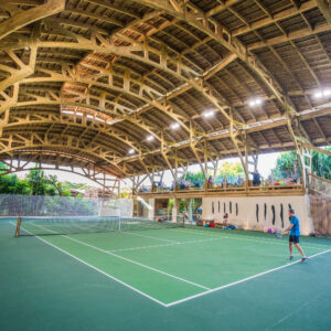 An indoor tennis court with a wooden arched ceiling. One player is on the court, ready to hit a ball, while several people sit on colorful bean bags to the left. The background features tropical plants and ample natural light filtering through the structure, reminiscent of Soneva Exclusive Offers&#039; tranquil settings.