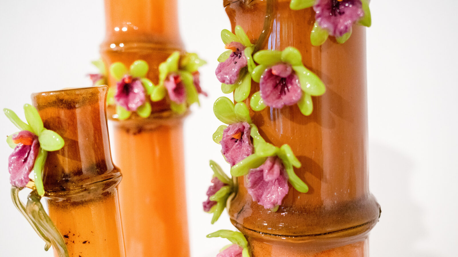 Close-up of decorative ceramic vases with an orange glaze, reminiscent of Mel Douglas at Soneva. The vases are adorned with intricate 3D floral details featuring purple and pink flowers with green leaves, all elegantly displayed against a plain white background.