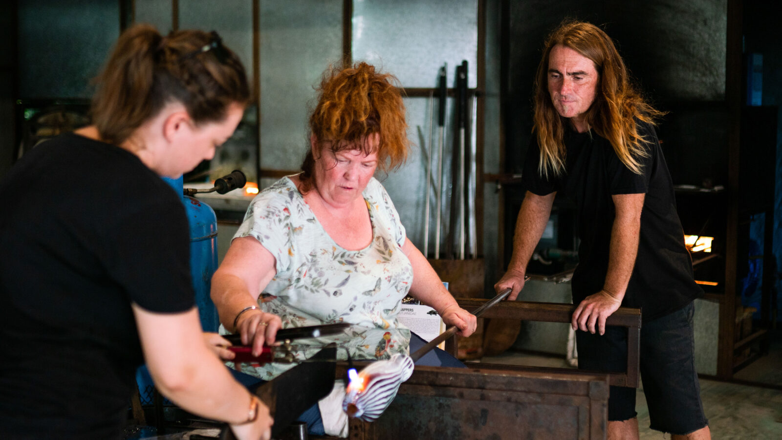 In a vibrant workshop filled with glassblowing tools, two individuals assist a red-haired woman in a floral shirt as she expertly shapes molten glass. Inspired by techniques from Mel Douglas at Soneva, the setting buzzes with creativity and craft.