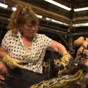 A person wearing gloves is carefully shaping a large, multicolored glass sculpture in a workshop reminiscent of Mel Douglas at Soneva. Glassblowing equipment surrounds them as another person assists, all under wooden beams and soft lighting.