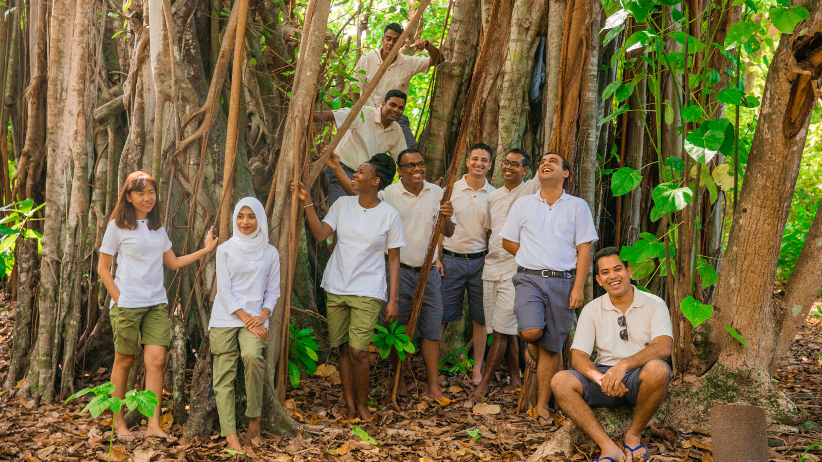 A group of nine people, dressed in white shirts and green shorts or khaki pants, stand happily among large tree roots in a forest. They are smiling and interacting with each other, enjoying the natural surroundings. Sunlight filters through the dense foliage, embodying the spirit of Soneva career opportunities.