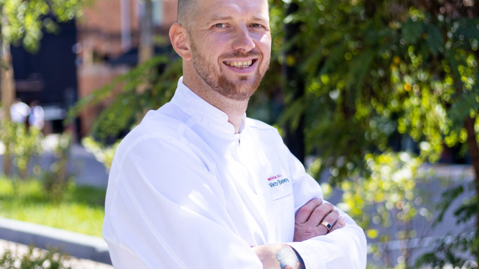 A person wearing a white chef's coat stands outdoors with arms crossed, smiling. They are in a garden setting with lush greenery in the background and sunlight filtering through the leaves.