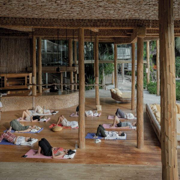 A group of people lie on yoga mats in various relaxing positions in a spacious, open-air wooden pavilion surrounded by lush greenery. A yoga instructor stands nearby, guiding the class. The setting features natural wooden elements and large windows providing an airy ambiance, reminiscent of a Soneva Award-winning resort in the Maldives.