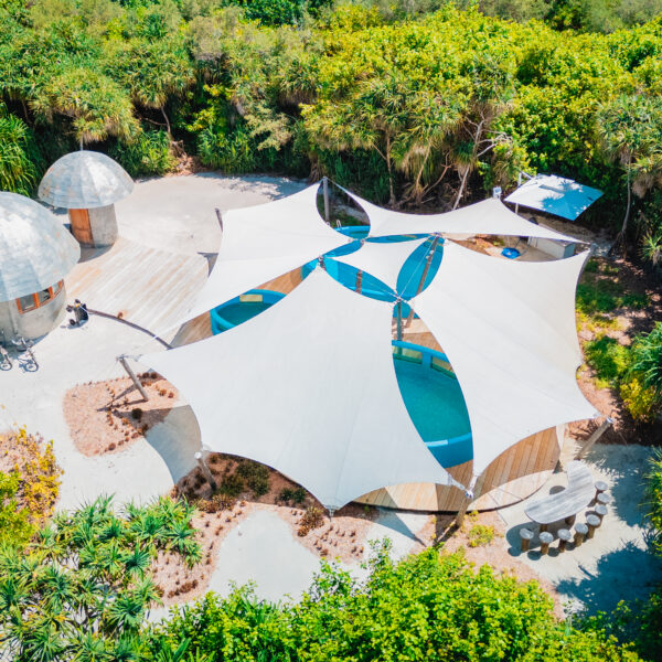 Aerial view of a unique architectural structure in a lush green setting, reminiscent of Soneva's style. The white tent-like roofs and circular wooden buildings are surrounded by dense foliage and trees. Nearby, a small patio with a table and bench from the Salt Group adds charm to the scenery.