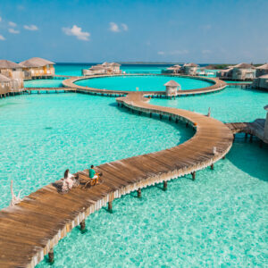 Aerial view of a tropical resort featuring wooden overwater villas connected by winding boardwalks. Two people are bicycling on the boardwalk over clear turquoise waters under a bright blue sky with a few clouds, enjoying their stay with Soneva Exclusive Offers.