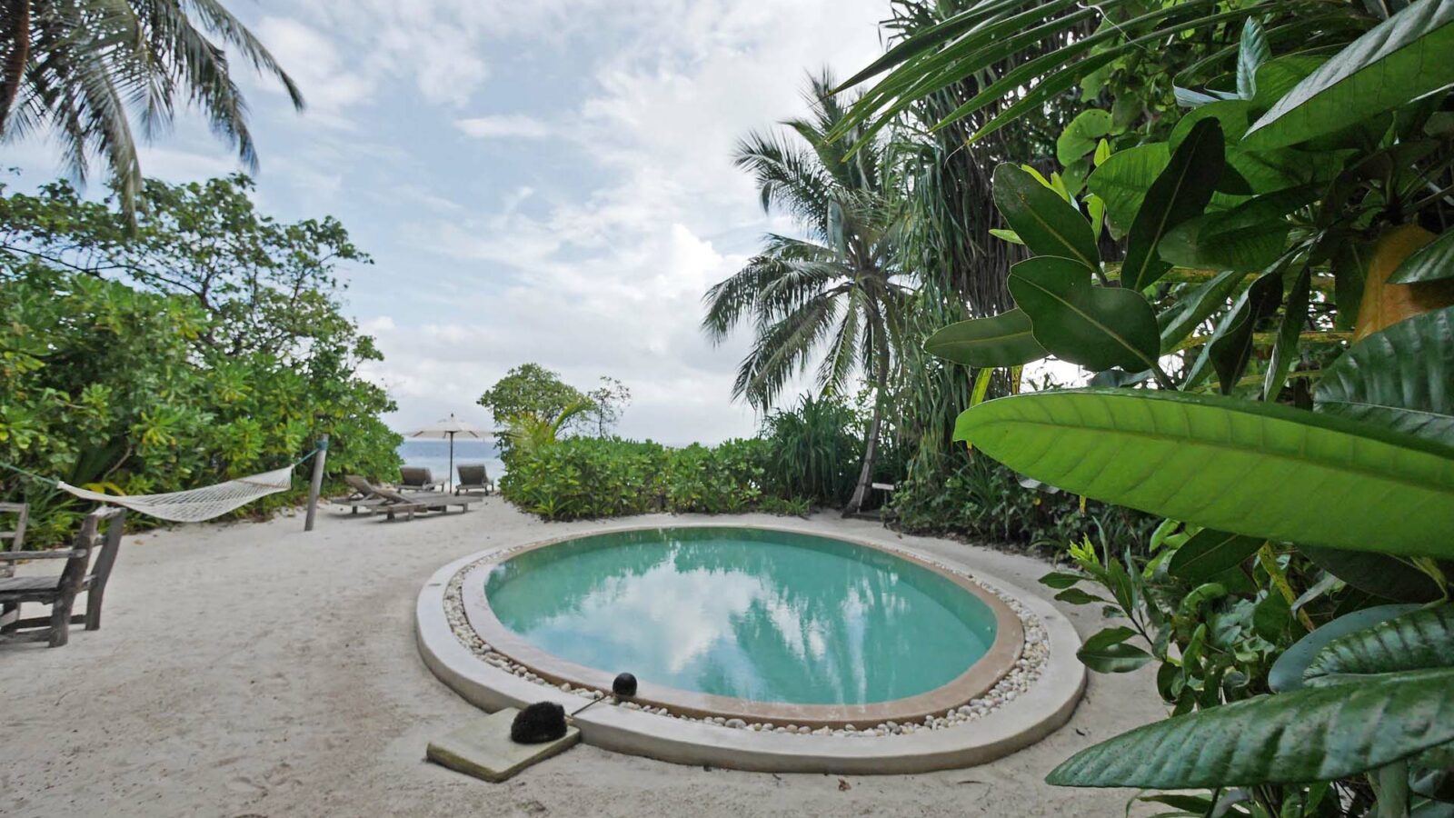 A small, round pool is surrounded by lush green plants and palm trees at the luxurious Soneva Fushi Villas in the Maldives. Nearby, a hammock is stretched between two trees. In the background, a pathway leads to a beach with blue ocean waves visible under a partly cloudy sky.