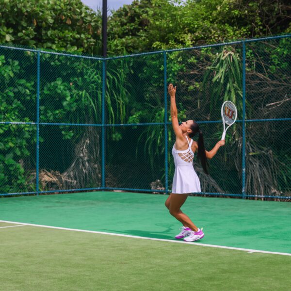 A woman in a white tennis dress prepares to serve on an outdoor tennis court surrounded by lush green foliage. With her arm extended upward and eyes focused on the ball, she embodies the elegance and precision of Soneva Exclusive Offers.