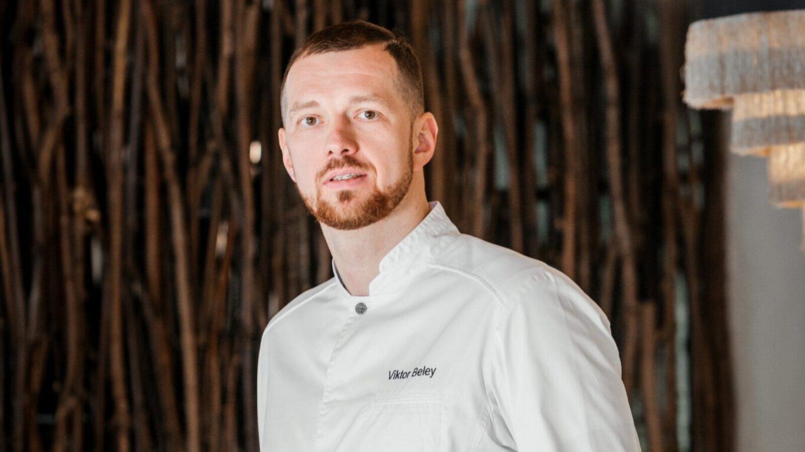 A man with a beard in a white chef's jacket stands confidently with hands in pockets. He is in a restaurant with modern decor, featuring a wooden backdrop and dining tables. The lighting is warm and ambient.