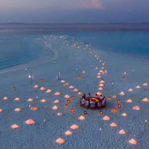 An aerial view of a beach at dusk reveals an enchanting atmosphere where a group of people is seated at a round dining table, surrounded by glowing lanterns and small tents. This magical scene could be part of the Soneva Exclusive Offers, creating unforgettable moments by the water.