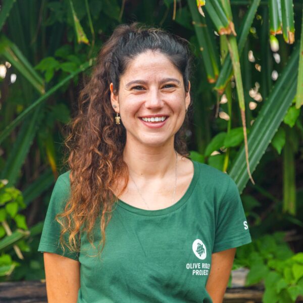 A person with long curly hair is smiling while standing in front of lush green foliage. They are wearing a green shirt with the text "Olive Ridley Project" on it.