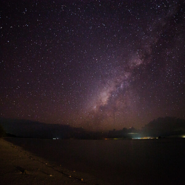 A serene night sky filled with stars and the Milky Way, seen over a calm Maldivian beach and ocean. The horizon shows distant lights, while the foreground includes a sandy beach and dark silhouettes of trees to the left, offering an exclusive Soneva experience.