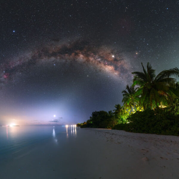 A serene tropical beach at night with palm trees and gentle waves. The sky is filled with stars, and the Milky Way galaxy arches across the horizon. Distant lights over the water add a soft glow to this Maldives exclusive experience, offering a glimpse of paradise at Soneva resorts.