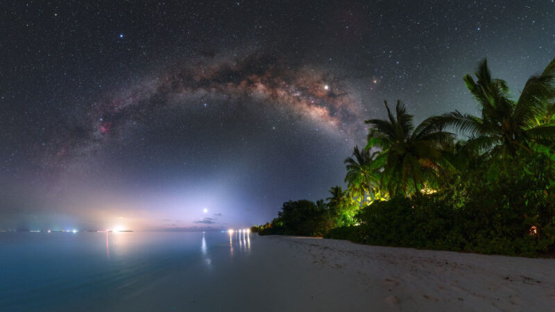 A serene tropical beach at night with palm trees and gentle waves. The sky is filled with stars, and the Milky Way galaxy arches across the horizon. Distant lights over the water add a soft glow to this Maldives exclusive experience, offering a glimpse of paradise at Soneva resorts.