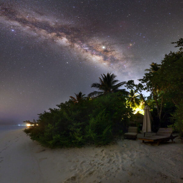 A stunning night view of a beach with clear skies showcasing the Milky Way galaxy. The sandy shore, lined with lush green trees and vegetation, offers an exclusive Maldives experience. Illuminated beach chairs and an umbrella nestled among the trees enhance this serene Soneva setting.