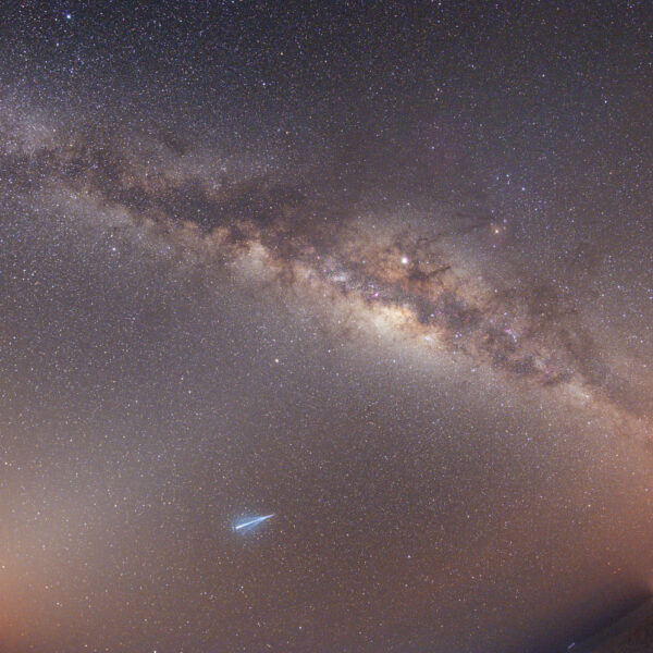 A striking image of the Milky Way galaxy sprawling across the night sky, with countless stars illuminating the scene. A shooting star is visible near the bottom, and a faint horizon with a few lights from buildings can be seen in the distance, creating an exclusive experience akin to a night in Soneva Maldives.