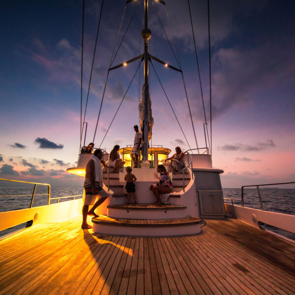 People gather on the deck of a yacht at sunset, with a few standing while others sit on steps. The sky is painted in hues of pink and purple, and the calm ocean reflects the serene beauty of a Maldives exclusive experience. Warm lights illuminate the deck, creating a relaxing atmosphere.