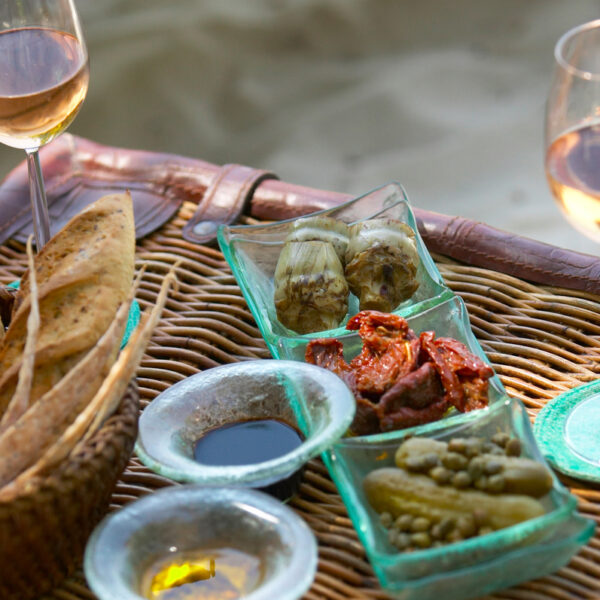 A wicker table displays a spread of assorted gourmet appetizers, including sun-dried tomatoes, pickles, and artichoke hearts in glass dishes. Two glasses of rosé wine are placed on the table alongside a basket of sliced bread, offering a taste of what one might savor during a Maldives Exclusive Experience at Soneva.