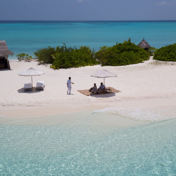 A beach with white sand and clear turquoise water. Two people are relaxing under a large beach umbrella with a table and drinks. A person in white is bringing them a drink. Thatched-roof huts and lush green bushes create the backdrop of this Maldives exclusive experience at Soneva.