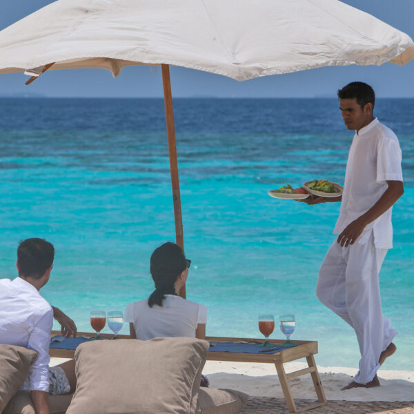 A couple sits at a beachside table under a white umbrella, enjoying drinks while a waiter brings a tray of food. The turquoise ocean and clear blue sky create a peaceful backdrop, epitomizing the Maldives Exclusive Experience at Soneva.