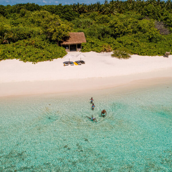 An aerial view shows a pristine white sandy beach with lush green vegetation in the background. Three people are snorkeling in the clear turquoise water near the shore. A small thatched-roof hut with lounge chairs is visible on the beach, capturing the essence of a Maldives Exclusive Experience.