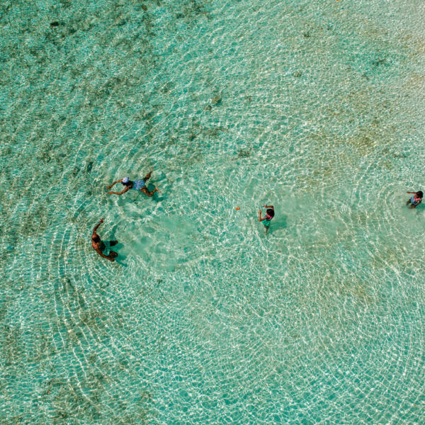 Aerial view of four people swimming and wading in a clear, shallow turquoise sea at Soneva. The water is calm, revealing the sandy bottom with patterns of light creating circular ripples around the people, encapsulating an exclusive Maldives experience.