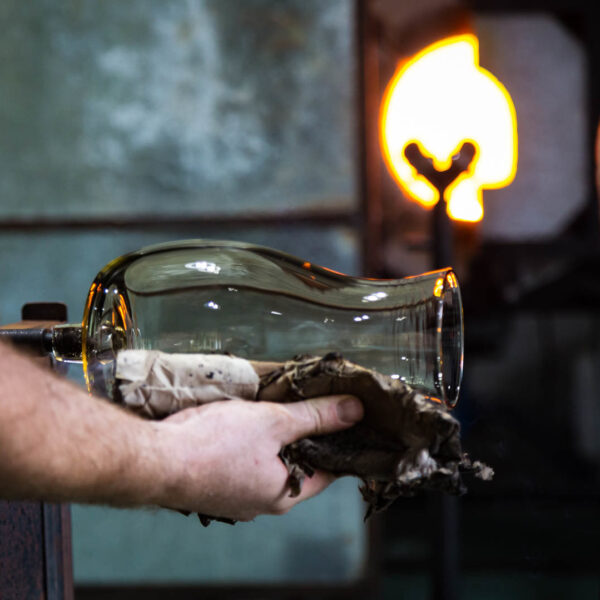 A glassblower carefully shapes a hot glass object with a wooden tool in front of a glowing furnace. The partially formed glass piece is being held by another person using a cloth for protection. The workshop, reminiscent of the exclusive experiences offered at Soneva in the Maldives, buzzes with heat and metallic elements.