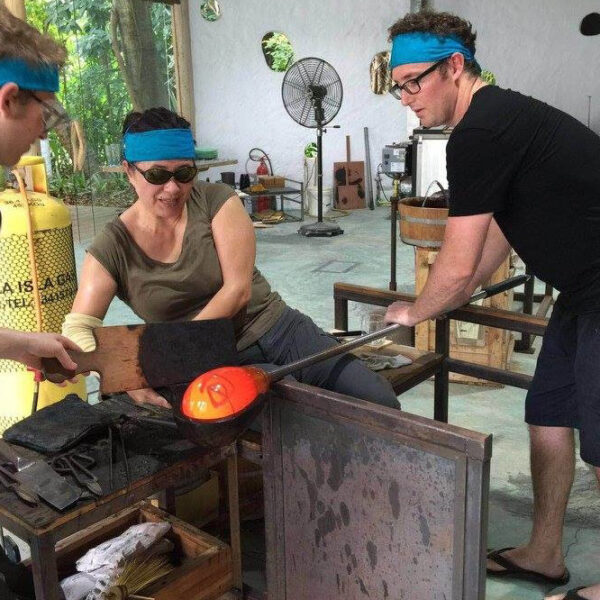 Three people, all wearing blue headbands and protective glasses, engage in glassblowing activities in a studio. One person guides a rod with molten glass while the others assist. The studio, reminiscent of a Maldives exclusive experience at Soneva, boasts various equipment and tools, including a large yellow gas cylinder.