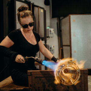 A woman in a black outfit and dark sunglasses is engaged in glassblowing, reminiscent of the meticulous artistry often seen at Soneva's Maldives Exclusive Experience. She uses a blowtorch to shape a glowing piece of glass amidst workshop equipment and a furnace.