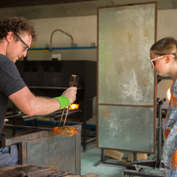 A male glassblower with curly hair demonstrates his craft to a young girl with braided hair and wearing a colorful dress. Using glassblowing tools, he creates intricate designs while she watches attentively, experiencing this Maldives exclusive experience at Soneva&#039;s renowned workshop.