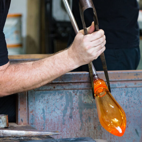 A glassblower is creating an orange glass object by shaping molten glass with a long metal tube. The artisan&#039;s gloved hands hold the tools carefully amid a workshop setting. The glowing glass piece, reminiscent of the brilliant sunsets in a Maldives exclusive experience at Soneva, contrasts with the metallic, rugged workbench surface.