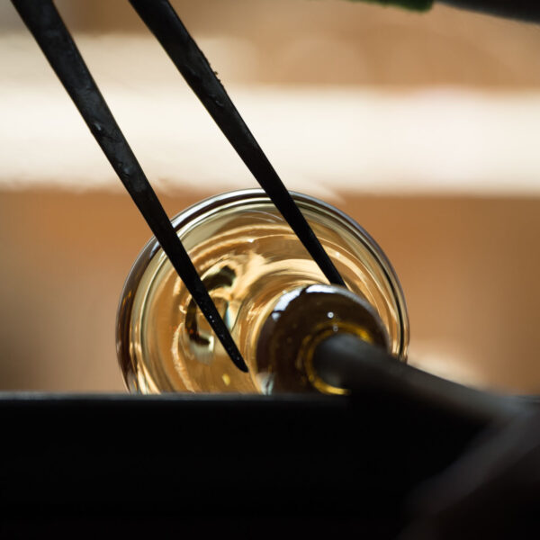 Close-up of a glassblowing process. A pair of pincers delicately shapes an incandescent sphere of glass at the end of a metal rod, glowing with a warm amber hue. The background is blurred, emphasizing the craftsmanship reminiscent of a Maldives Exclusive Experience at Soneva resorts.