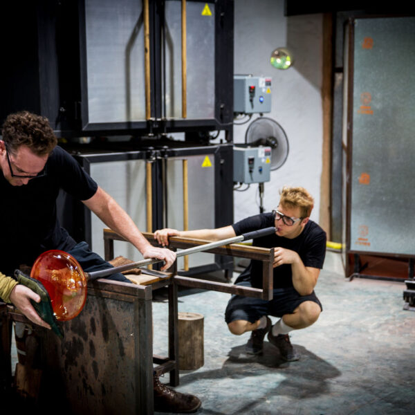 Two people in a glassblowing studio work together on shaping molten glass. One person manipulates the glass with tools, while the other assists with a long rod. They wear protective eyewear and gloves as the furnace glows intensely in the background, crafting what could be a Maldives exclusive experience at Soneva.