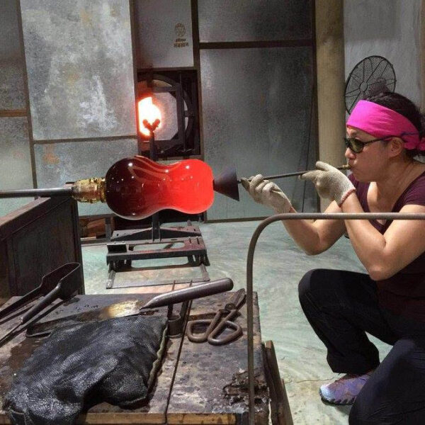 A person wearing a pink headband and safety glasses is shaping molten glass with a blowpipe in a glassblowing studio. The red-hot glass piece is being heated in a furnace while various tools are placed on a table nearby, making it feel like an exclusive experience at Soneva in the Maldives.