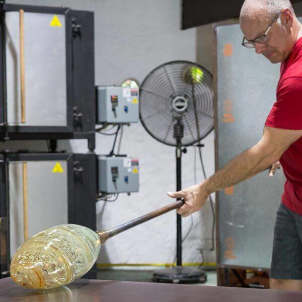 A person wearing a red shirt and glasses is shaping hot glass using a blowpipe in a glassblowing workshop. Industrial equipment, including a fan and control panels, are visible in the background. The partially-formed glass piece is glowing and semi-transparent, an artistry reminiscent of a Maldives Exclusive Experience at Soneva.