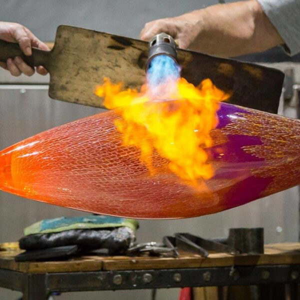 Two people use a handheld torch and wooden paddles to shape a large, glowing piece of molten glass in a workshop. The glass is orange with purple accents, and flames are visible from the torch. Tools and equipment are scattered on a workbench nearby—a true Maldives Exclusive Experience at Soneva.