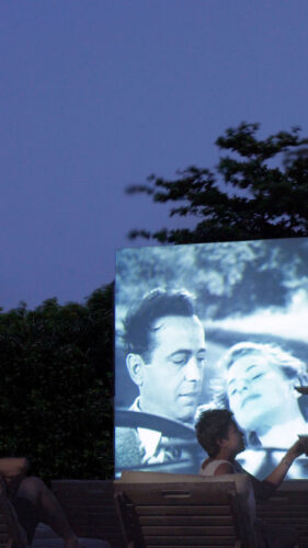 Under a moonlit sky, two people relax on lounge chairs, indulging in an exclusive Soneva experience while watching an outdoor movie projected onto a screen. The black-and-white film depicts a romantic scene. A waiter in white uniform serves drinks to one of the viewers. Trees and bushes frame the background.