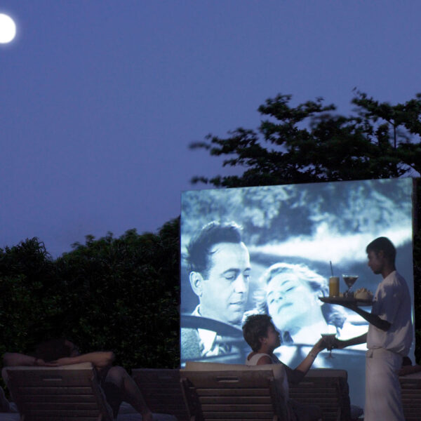 Under a moonlit sky, two people relax on lounge chairs, indulging in an exclusive Soneva experience while watching an outdoor movie projected onto a screen. The black-and-white film depicts a romantic scene. A waiter in white uniform serves drinks to one of the viewers. Trees and bushes frame the background.