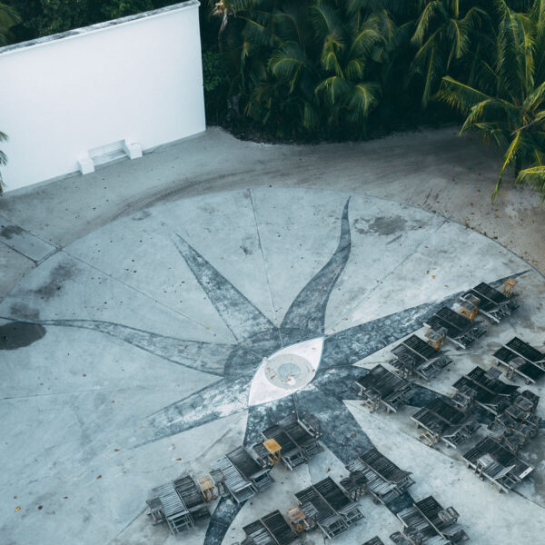 Aerial view of an exclusive outdoor cinema surrounded by lush palm trees at Soneva in the Maldives. The ground is decorated with a large, star-like pattern, and several rows of lounge chairs face a white projection screen. The area appears quiet and serene.