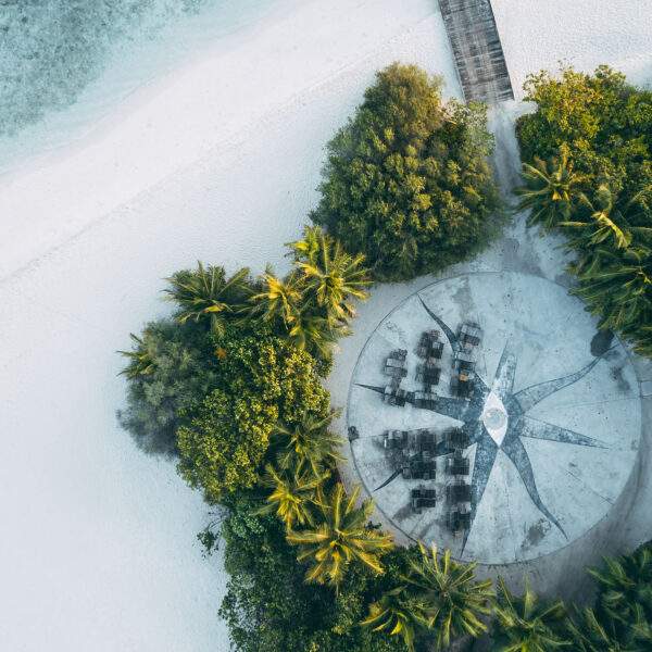 Aerial view of a circular beachside seating area surrounded by lush greenery and palm trees. The white sandy beach and turquoise waters are visible to the left, with a wooden pathway leading to the seating area from the top right corner—an exclusive Maldives experience reminiscent of Soneva's luxurious retreats.