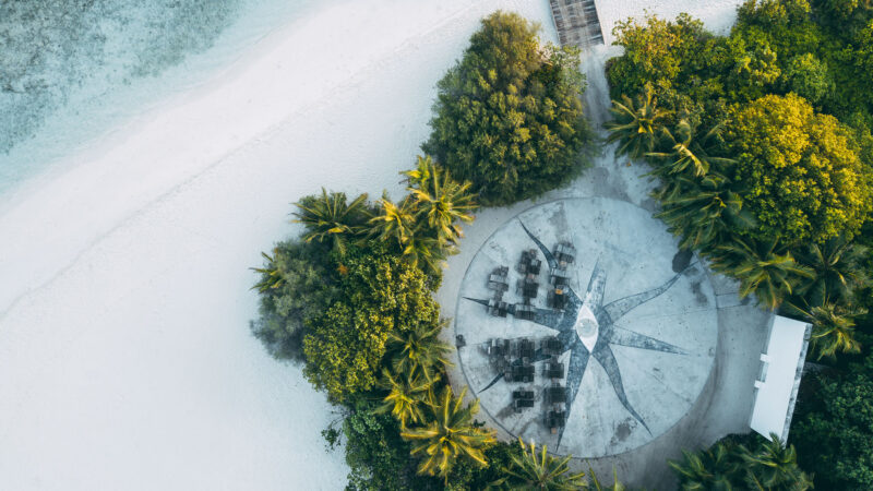 Aerial view of a circular beachside seating area surrounded by lush greenery and palm trees. The white sandy beach and turquoise waters are visible to the left, with a wooden pathway leading to the seating area from the top right corner—an exclusive Maldives experience reminiscent of Soneva's luxurious retreats.