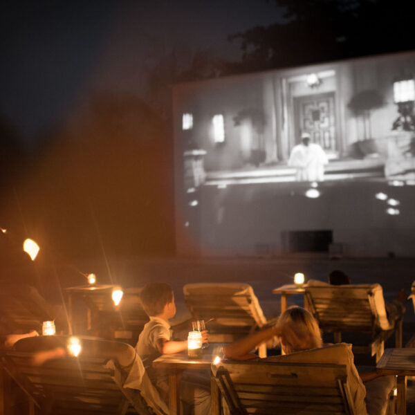 A group of people sit in lounge chairs in an outdoor setting at night, watching a black-and-white movie projected on a large screen. Small lanterns on tables beside them provide a warm, ambient glow, creating an exclusive experience reminiscent of the serene luxury found at Soneva in the Maldives.