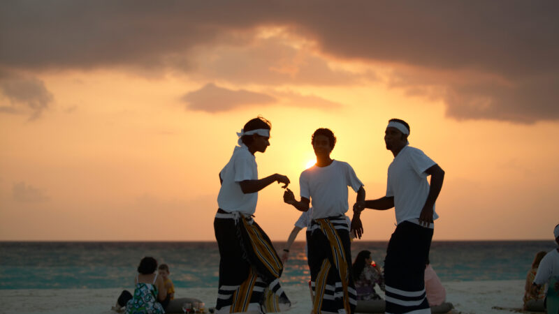 Three people dressed in white shirts and striped sarongs dance on a beach at sunset during the Maldives Exclusive Experience by Soneva. The sky is a blend of orange and pink, with a few people sitting near the water. The dancers' silhouettes are beautifully illuminated by the setting sun.